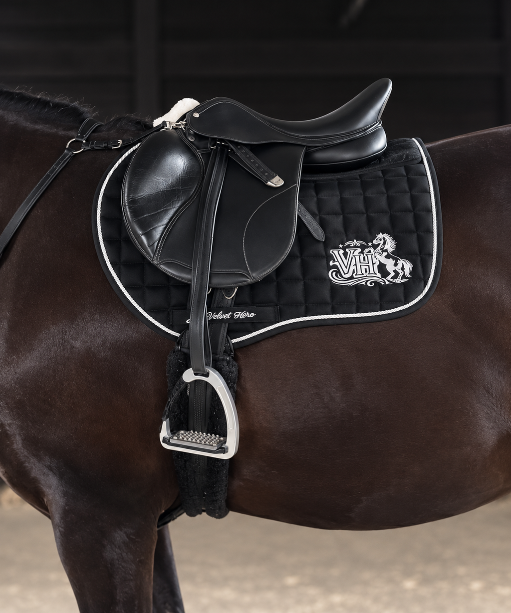 A close-up of a horse wearing a black leather western saddle with a black quilted saddle pad featuring white trim and a white embroidered logo.