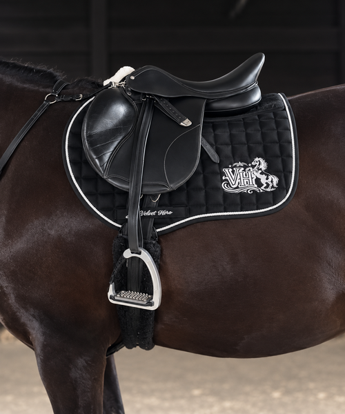 A close-up of a horse wearing a black leather western saddle with a black quilted saddle pad featuring white trim and a white embroidered logo.